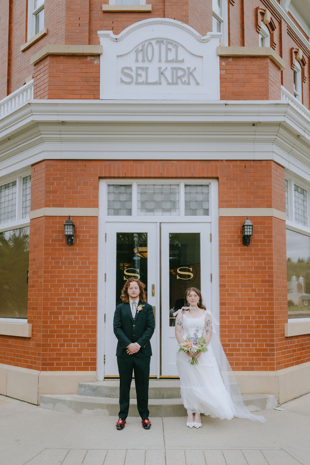 Bride and groom stand apart in front of Hotel Selkirk brick building on their wedding day.