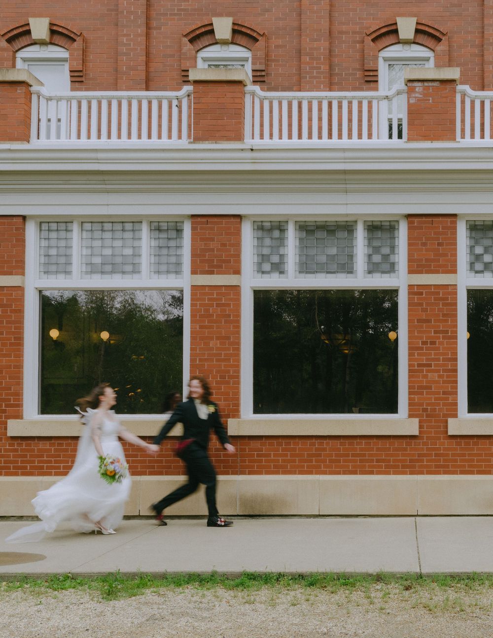 Bride and groom joyfully running hand-in-hand outside a classic red brick building on their wedding day.