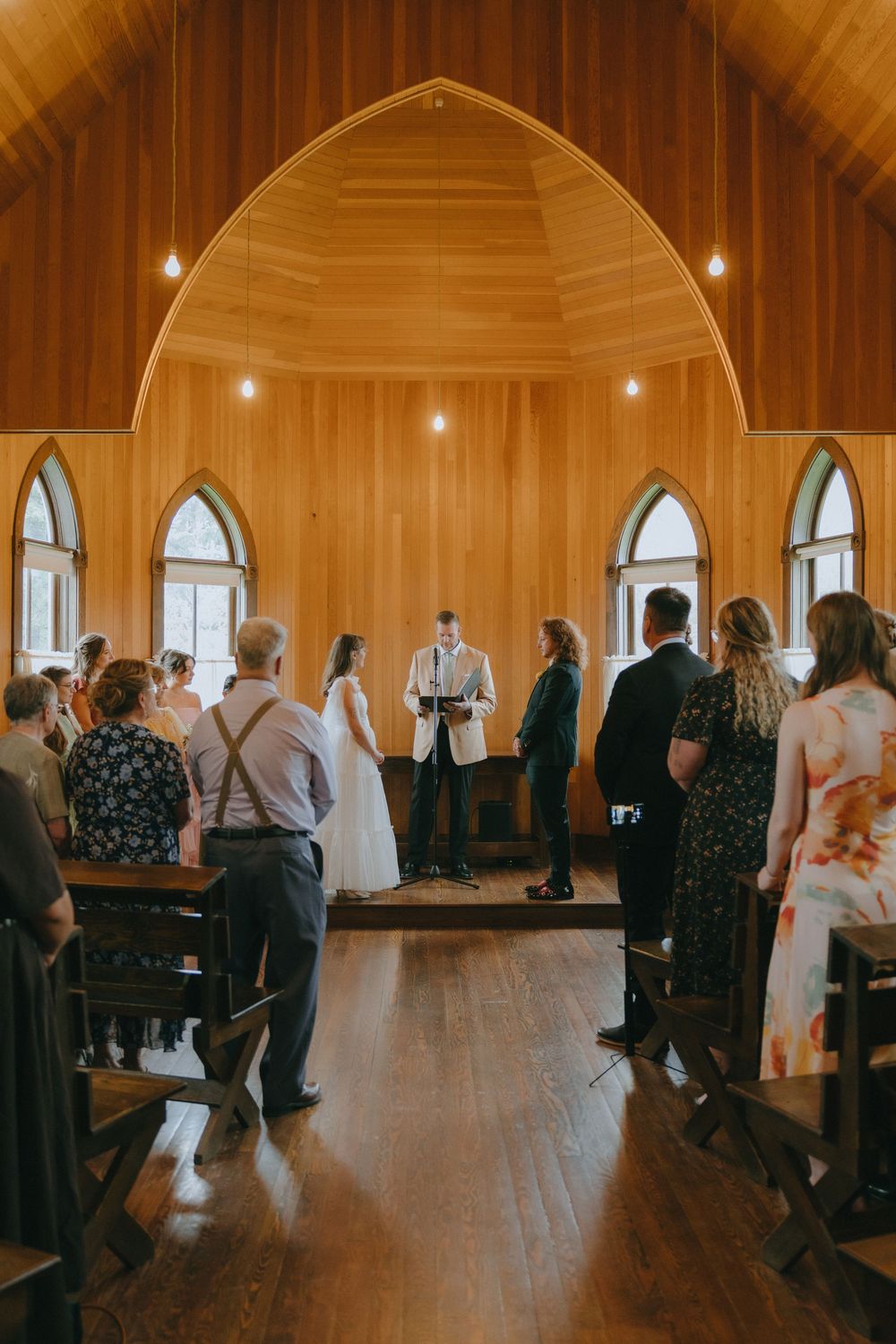 A wedding ceremony inside a wooden chapel with guests standing in pews as the couple exchanges vows at the altar.