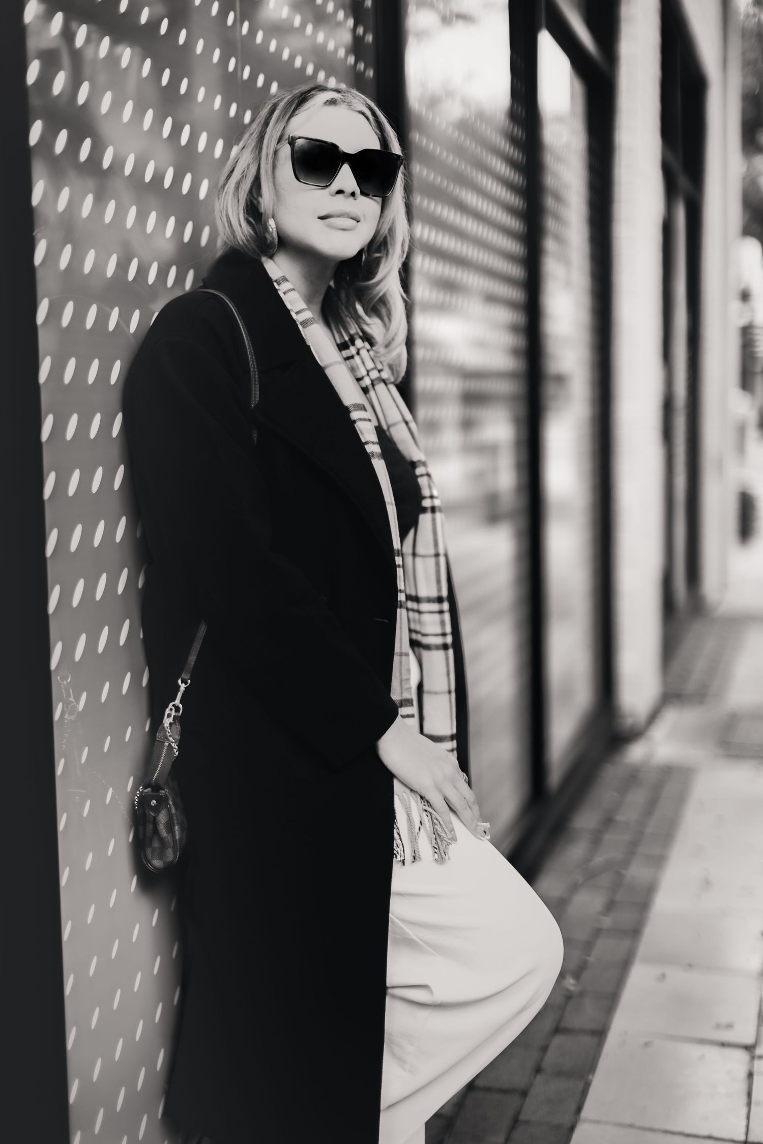 Black and white photo of a stylish blonde woman in sunglasses, long black coat, and plaid scarf leaning against a wall.