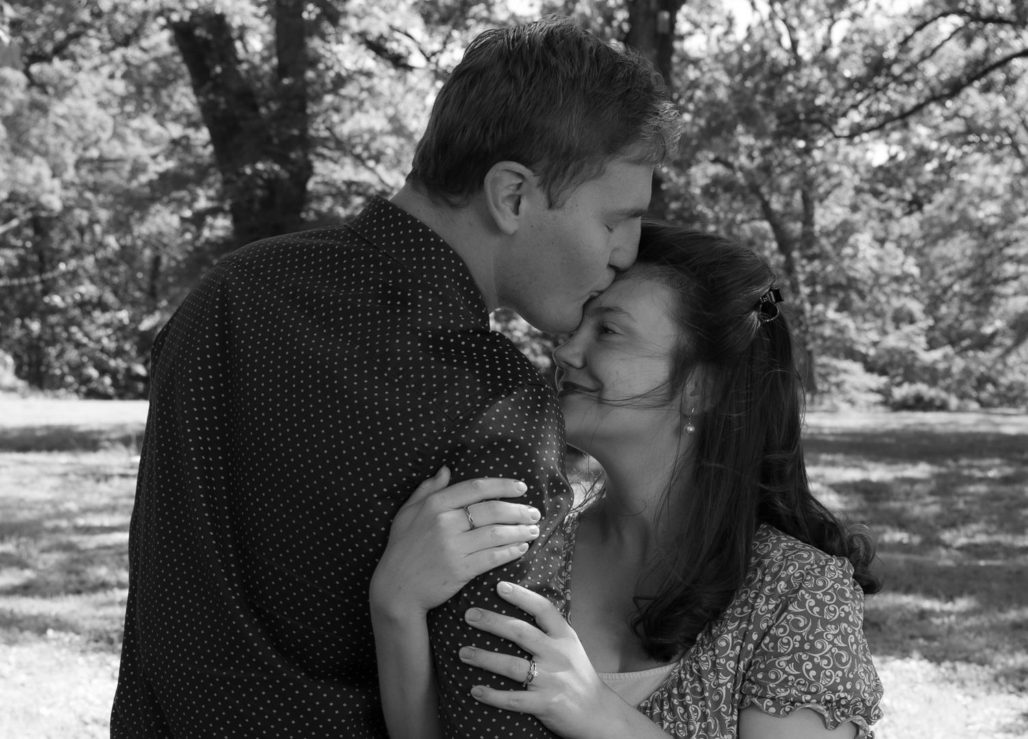 A couple sharing a romantic kiss outdoors in a black and white photo surrounded by lush trees.