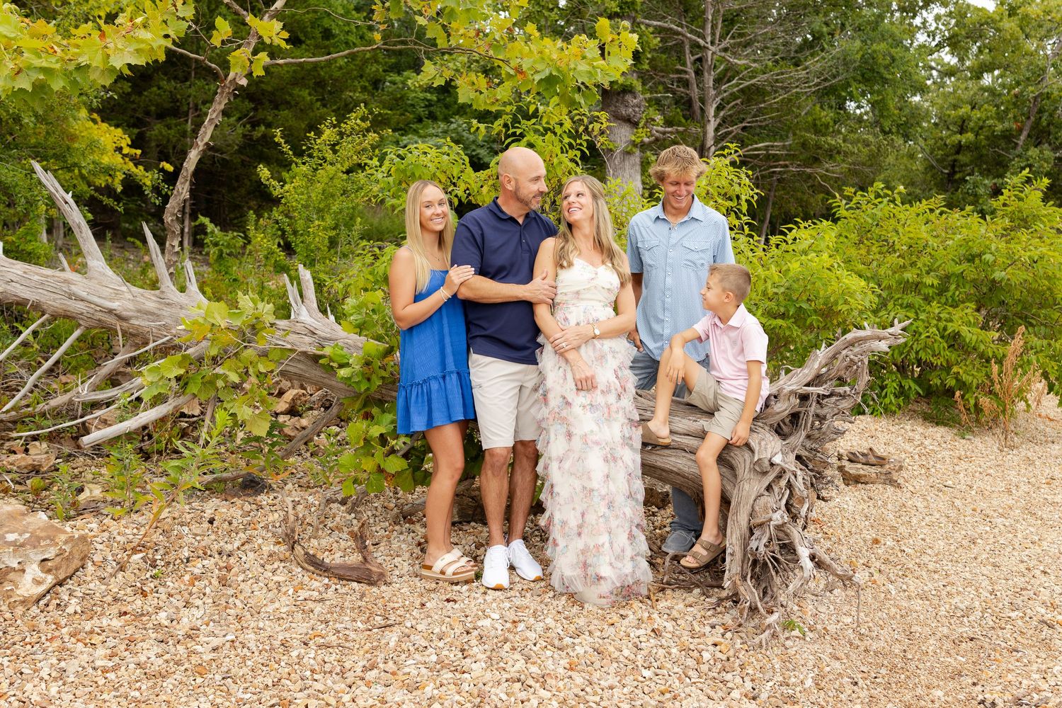Family of five posing outdoors near driftwood logs surrounded by lush green trees on a sunny day.
