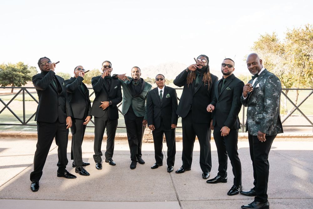 A group of eight groomsmen in black suits pose together outdoors at a wedding celebration.