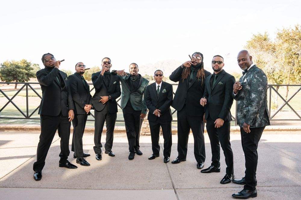 Groom and groomsmen in black suits and sunglasses posing outdoors at a wedding celebration.
