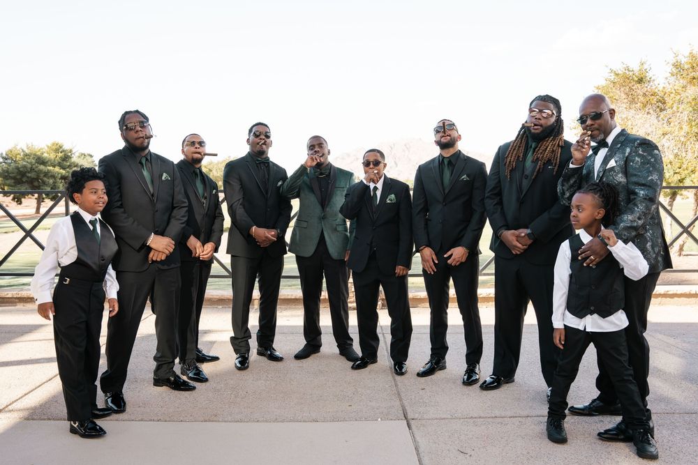Groomsmen in black suits with green accents pose with groom in teal jacket at outdoor wedding celebration.