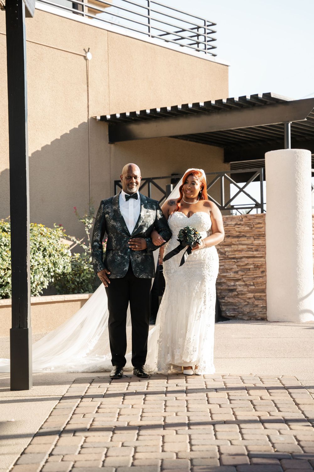 A smiling bride in a white lace gown holds a bouquet beside a dapper groom in a floral tuxedo jacket outdoors.