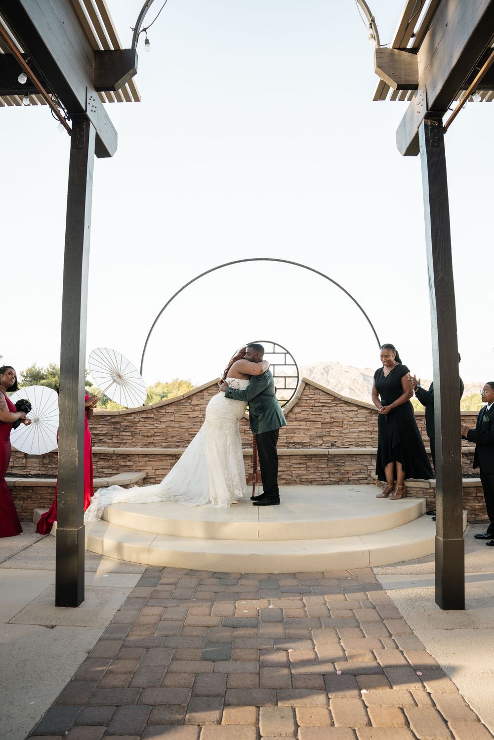 Bride and groom share first kiss on elevated platform under circular arch at outdoor desert wedding ceremony.