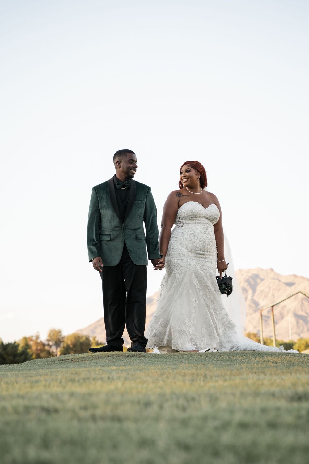 A smiling bride and groom hold hands on a grassy hill at sunset, she in a white gown, he in a green velvet suit.