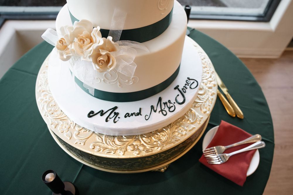 Elegant two-tier white wedding cake with green ribbon, gold floral details, and Mr. and Mrs. Jones inscription on stand.