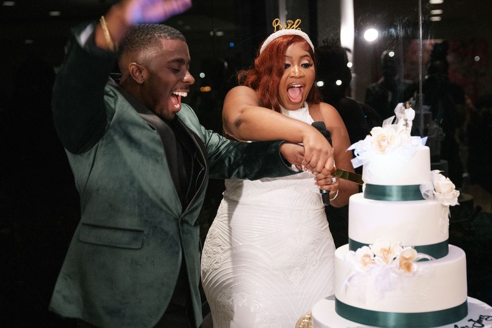 Joyful couple cutting white four-tier wedding cake, bride in gown with crown, groom in green suit.
