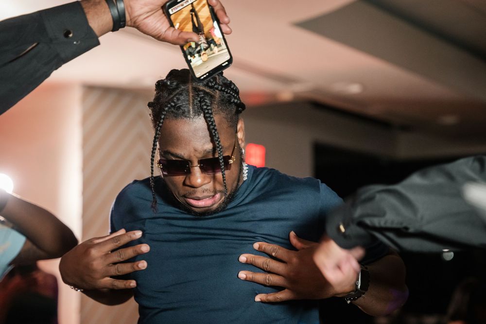 Man with braids and sunglasses reacts at an indoor event while someone holds a phone above his head.