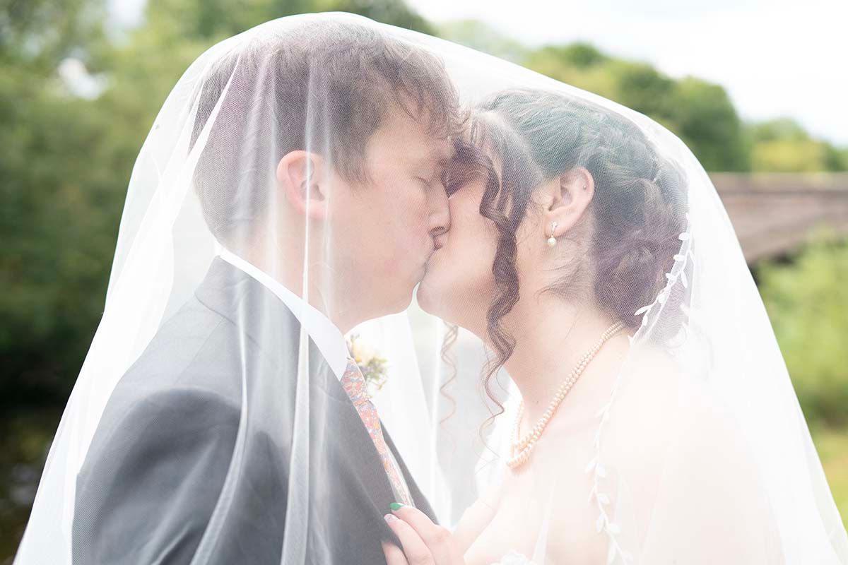 Bride and groom sharing a romantic kiss under a white wedding veil outdoors on their wedding day.