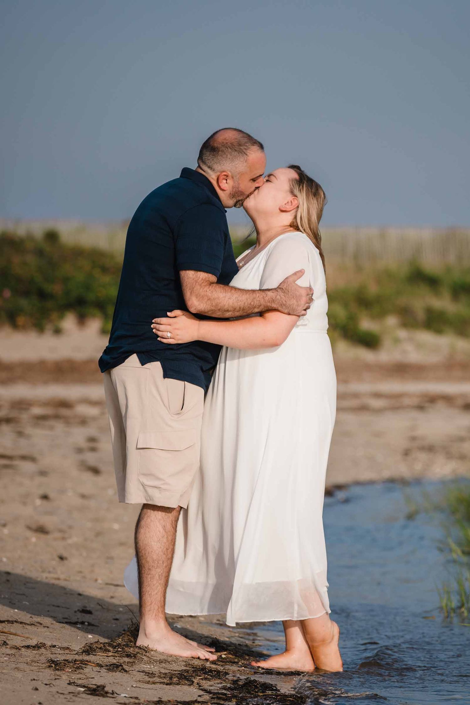 Engaged couple kissing barefoot in the shallow water at Duxbury Beach, Massachusetts, during golden hour light.