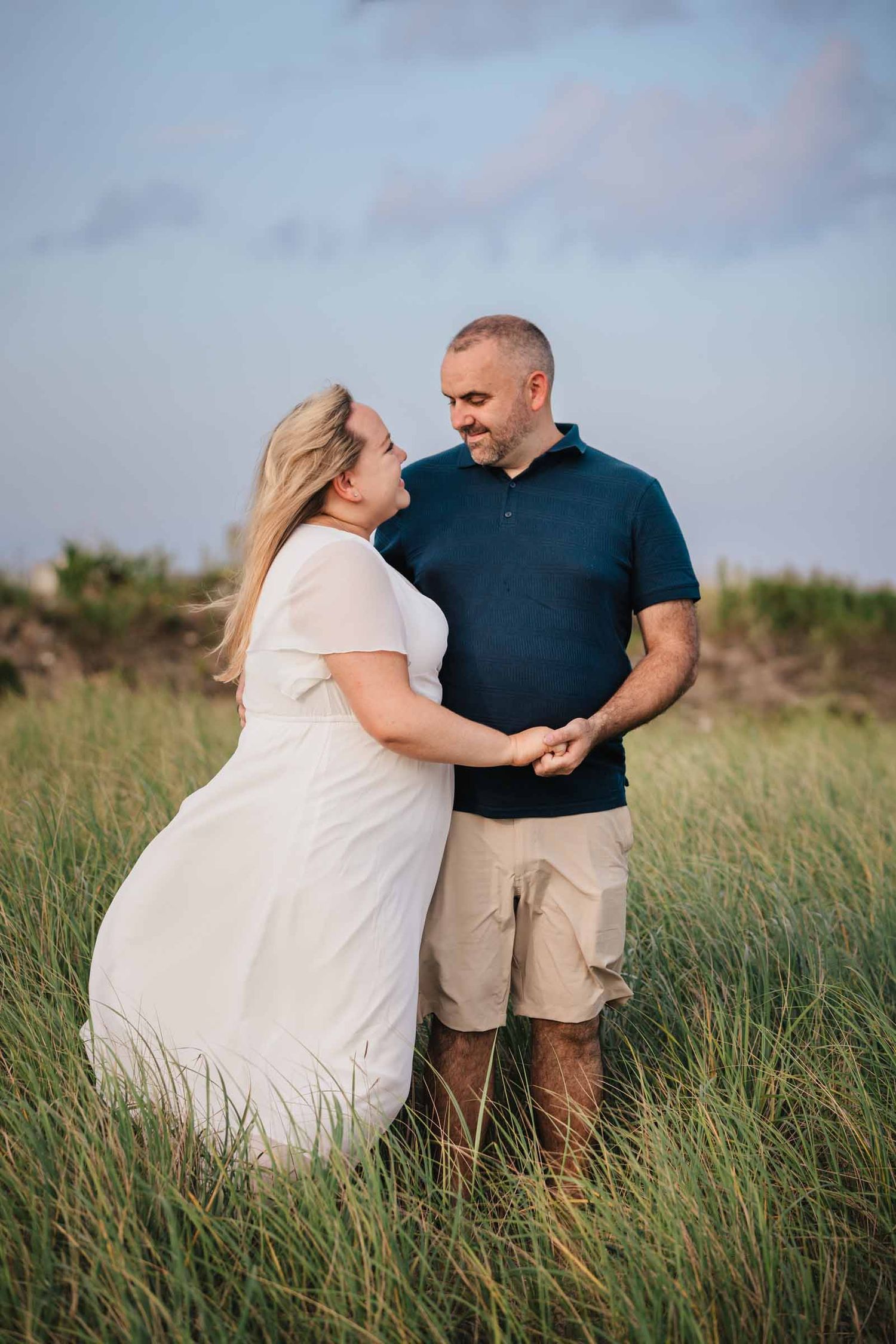 Engaged couple holding hands and laughing in tall grass at Duxbury Beach, Massachusetts, her dress caught by the wind