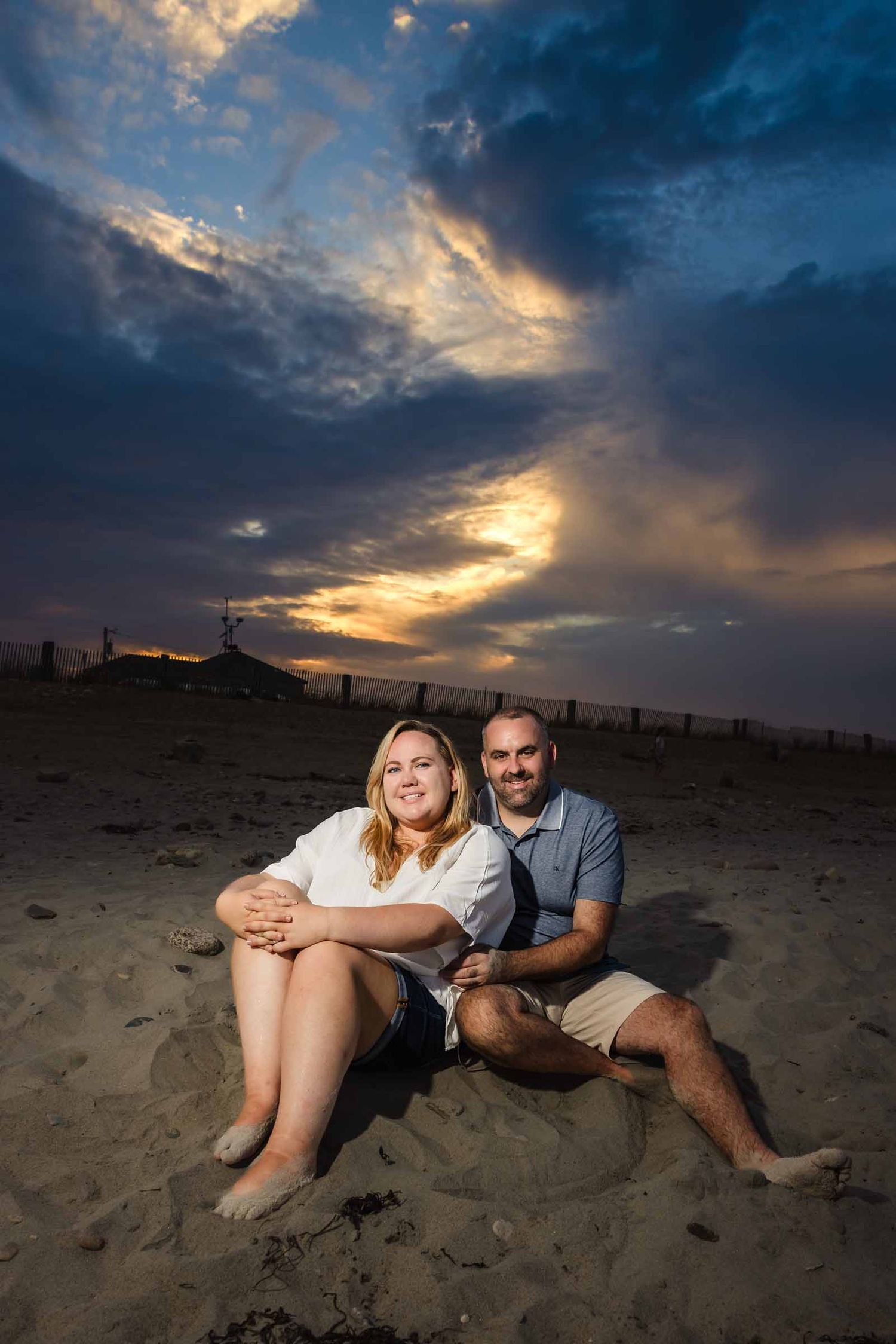 Couple sitting on sandy beach at sunset with dramatic cloudy sky in background.