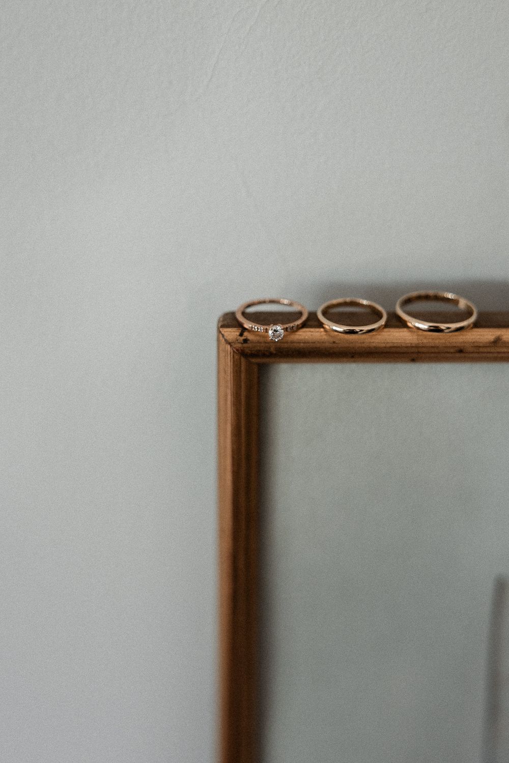 Three gold rings displayed on top of a wooden picture frame against a soft gray wall.