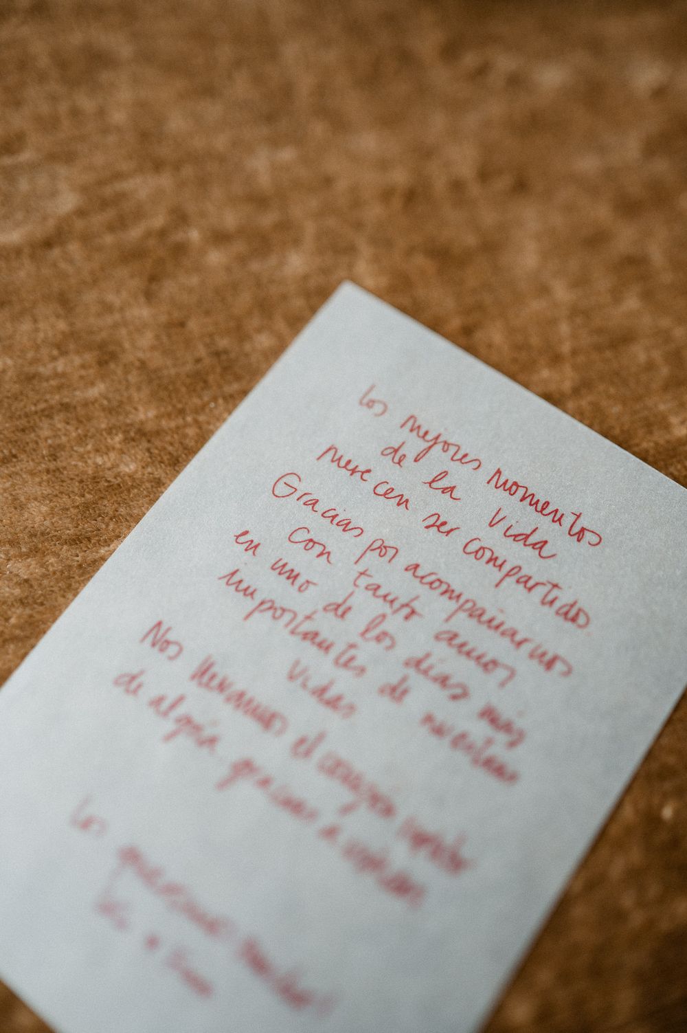 Handwritten note in Spanish with red ink on white card, placed on brown velvet surface at a wedding.