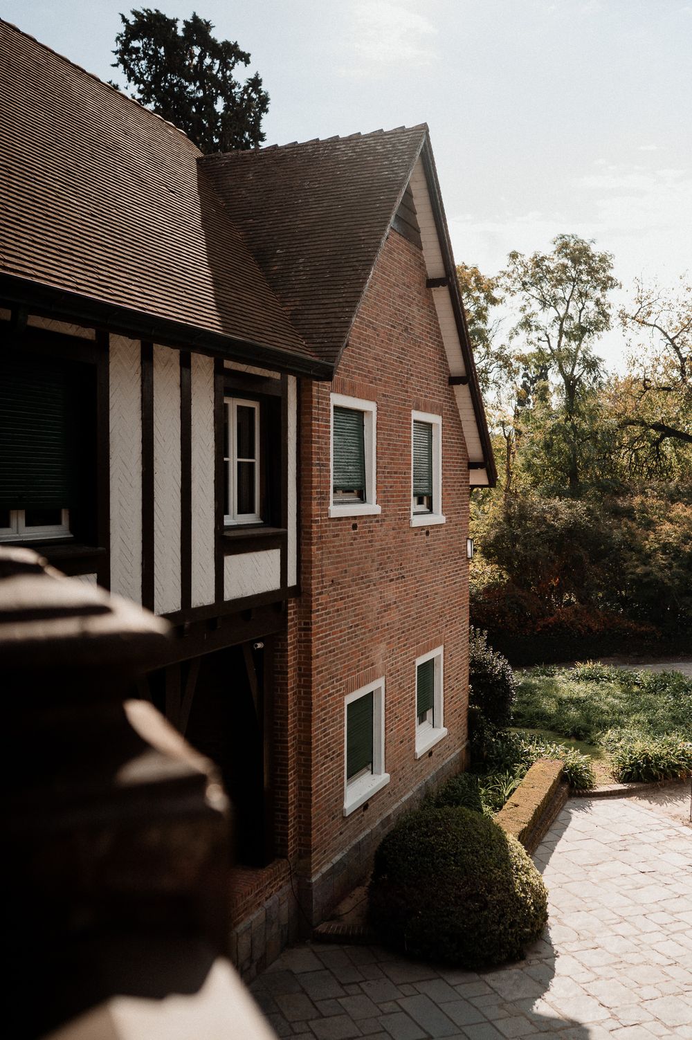 Charming brick and half-timbered house with manicured hedges surrounded by lush trees on a sunny day.
