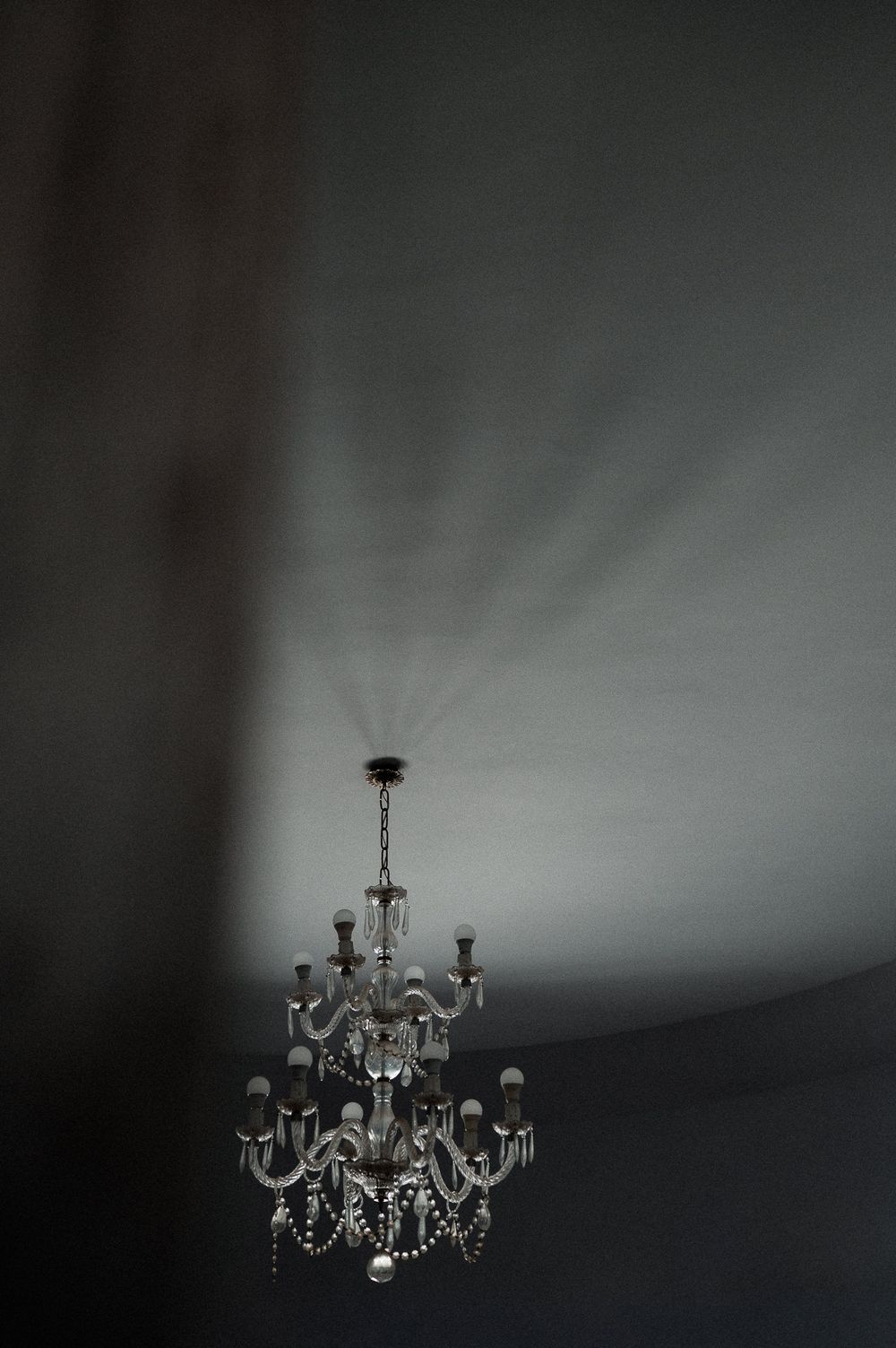 Crystal chandelier hanging from ceiling casting dramatic light rays against dark gray walls in dimly lit room.