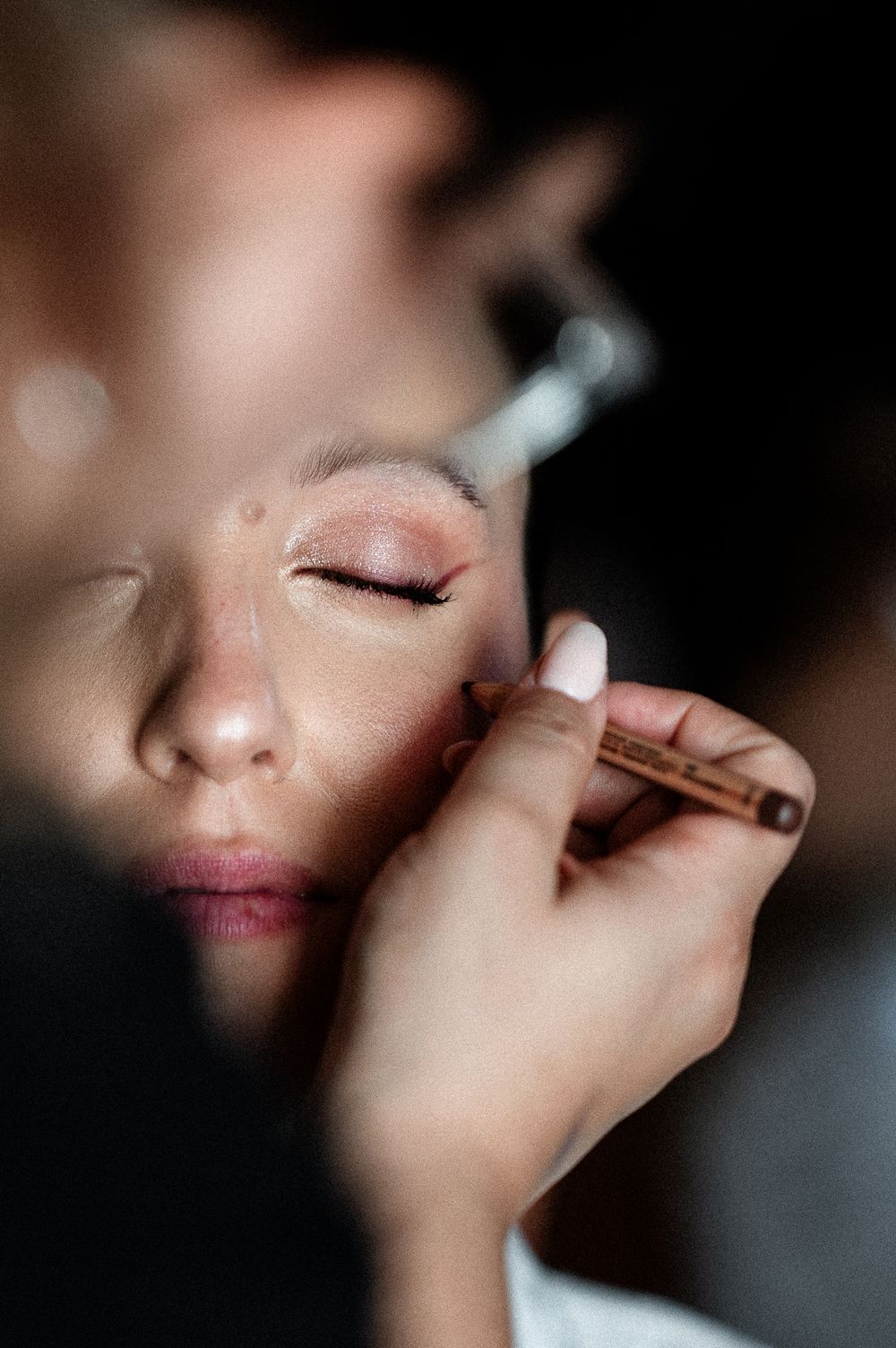 A makeup artist applies eyeliner to a model with closed eyes using a precise brush in a close-up beauty shot.