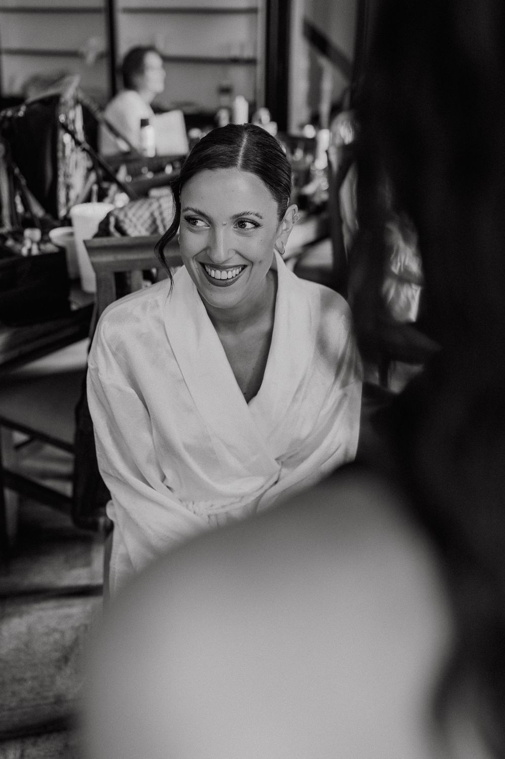 Radiant bride in white robe smiling joyfully during wedding preparations in black and white photo.