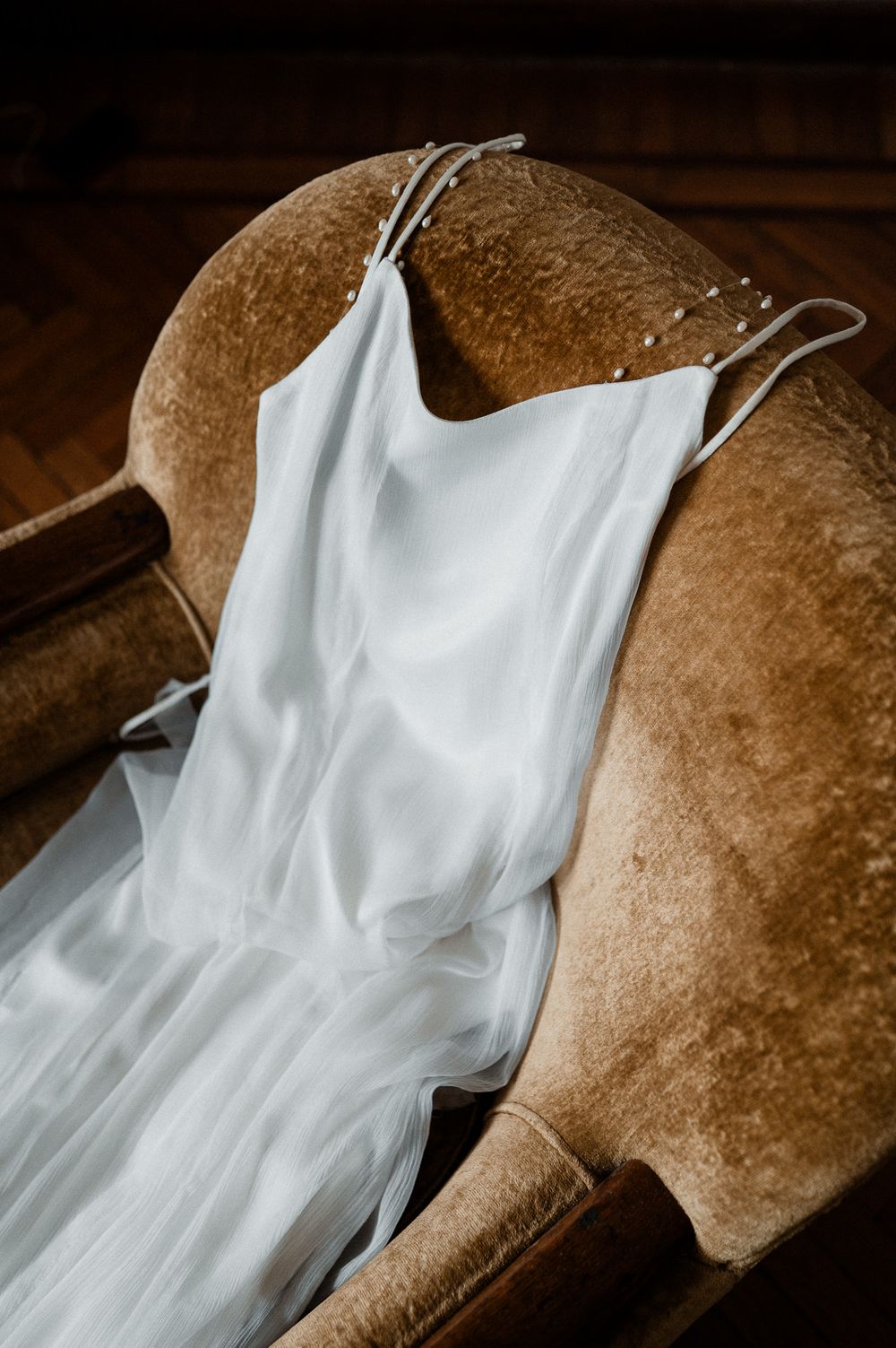Delicate white spaghetti strap wedding dress with sheer tulle overlay draped over a vintage velvet armchair.