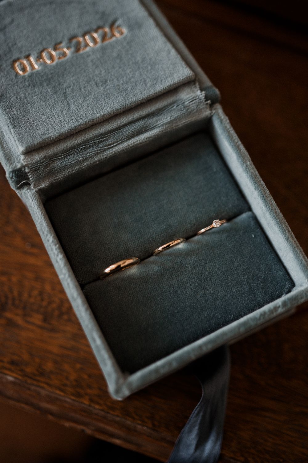 Two rose gold wedding bands in a gray velvet ring box embossed with date 01.05.2026 on dark wood.