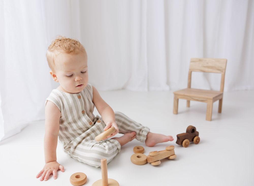 Baby boy sitting on floor playing with wooden toys in a bright white room wearing striped romper.