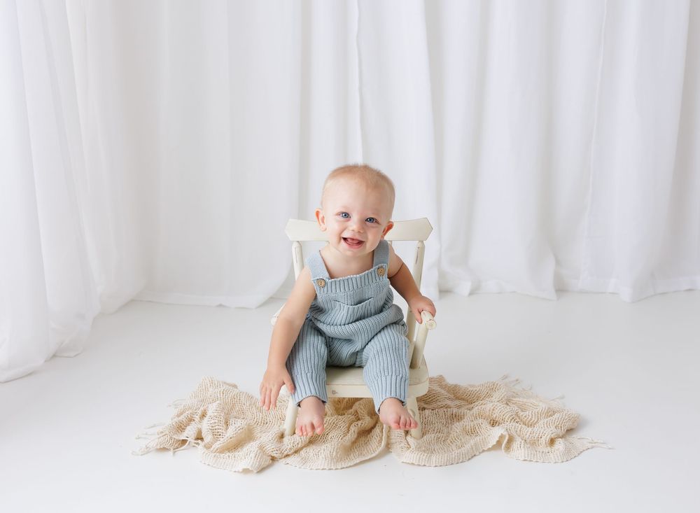 Smiling baby in blue knit romper sitting on white chair with beige blanket in bright white studio.