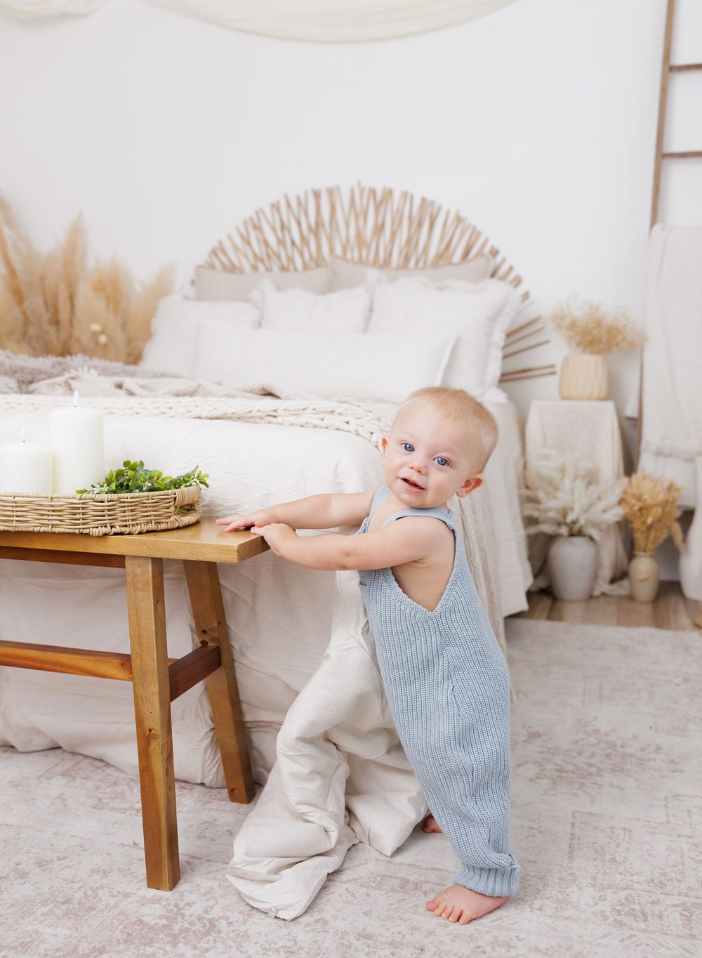 A smiling baby in a blue knit romper stands at a wooden table in a bright boho bedroom with neutral decor.