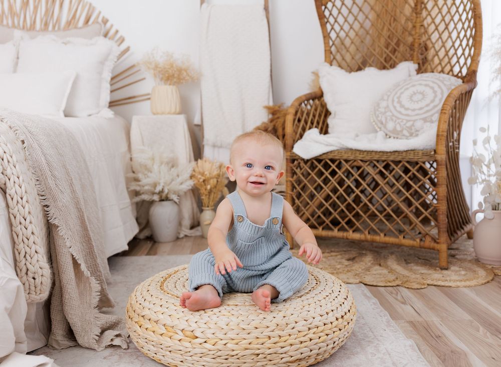 Smiling baby in gray overalls sitting on a woven pouf in a bright boho nursery with rattan chair.