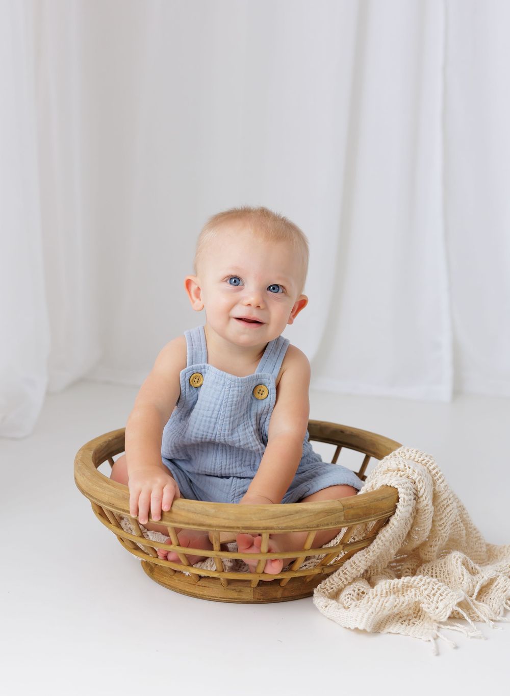 Smiling baby with blue eyes sitting in a wicker basket wearing a blue romper against a white background.