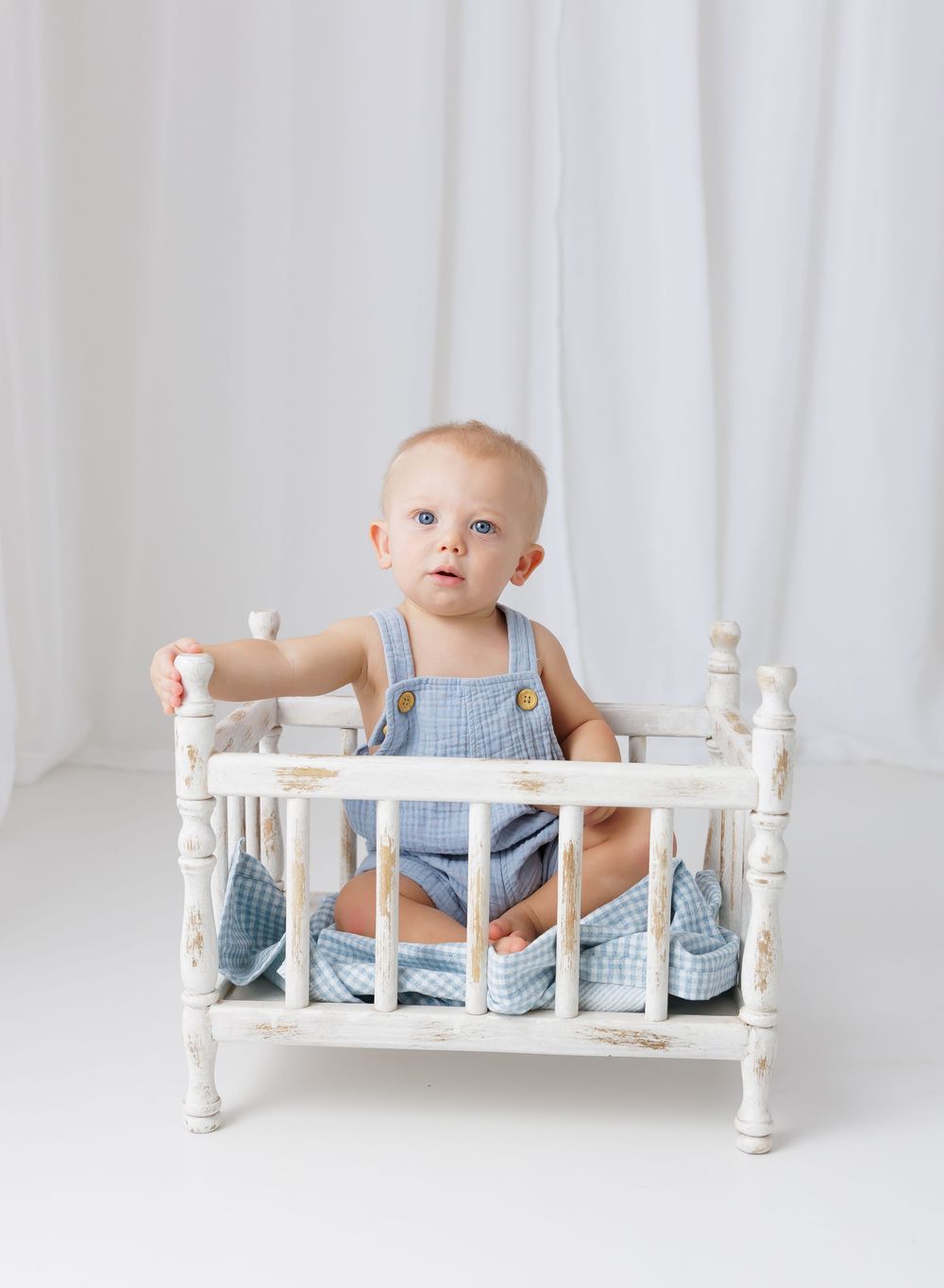 Baby boy in blue gingham overalls sits in a vintage white distressed doll crib against a white backdrop.