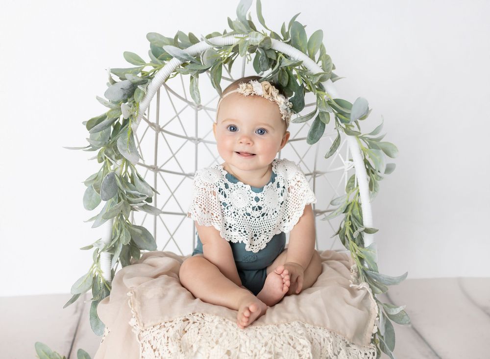 Smiling baby girl in white lace outfit sits in a wicker chair decorated with greenery for a professional photo session.