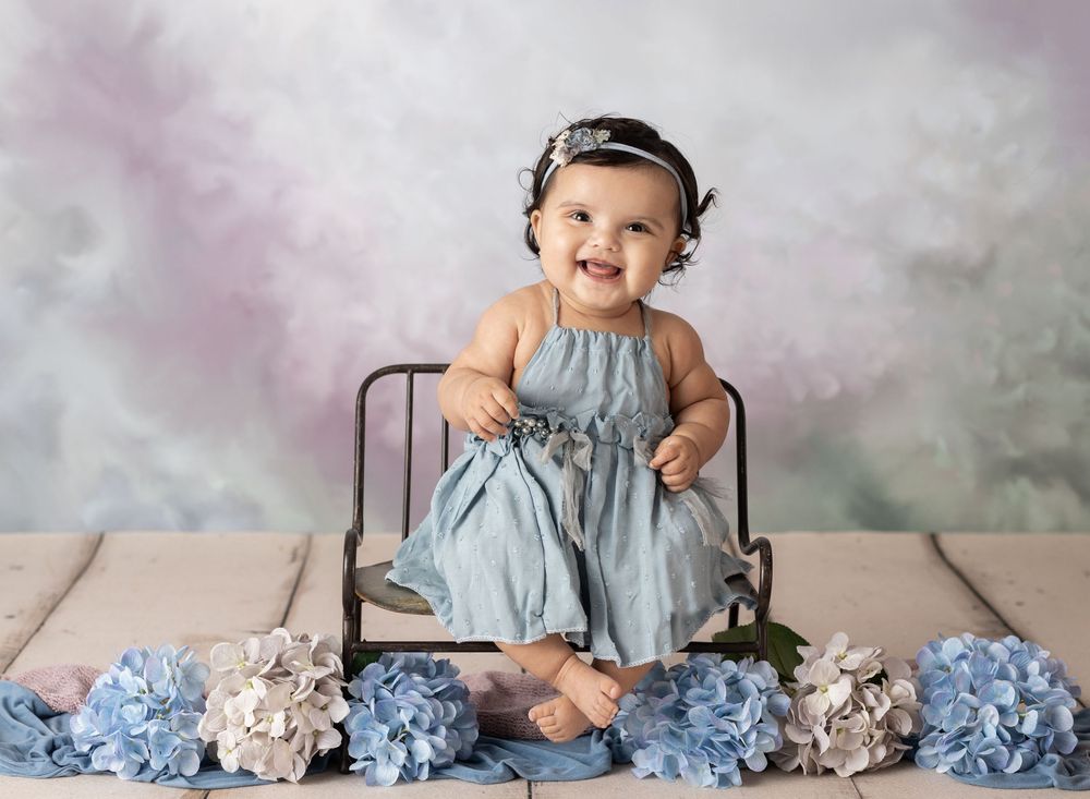 Smiling baby girl in blue dress sitting on vintage chair surrounded by blue hydrangeas against painted backdrop.