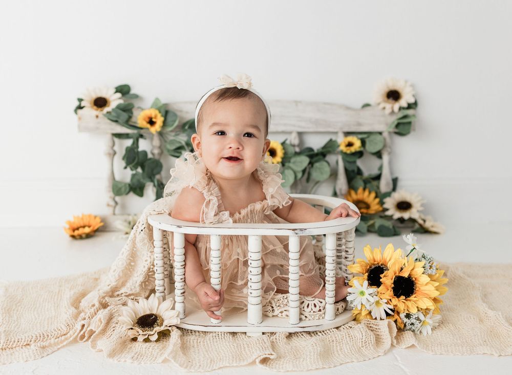 Smiling baby girl in lace romper sitting in white basket crib surrounded by sunflowers on white backdrop.