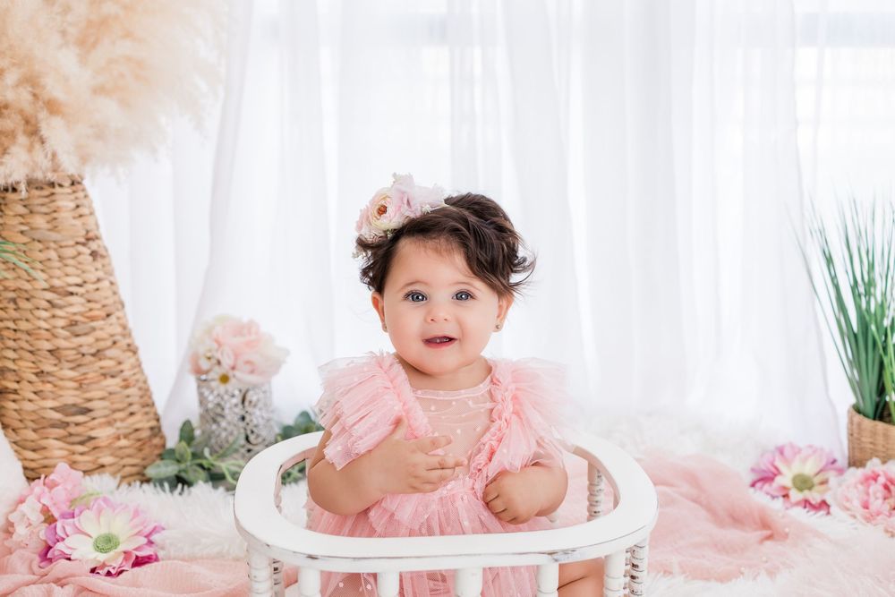 A cute baby girl in a pink ruffle dress and floral headband sits in a white chair surrounded by pink flowers.