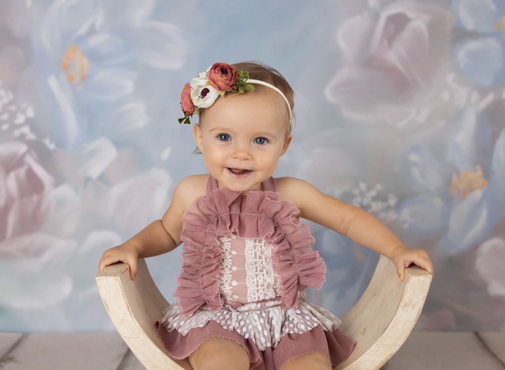 Smiling baby girl in pink ruffle romper with floral headband sits in wooden bowl chair against floral backdrop.