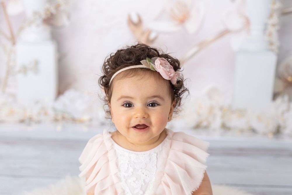 Smiling baby girl in pink ruffled dress and floral headband poses for a professional first birthday portrait.
