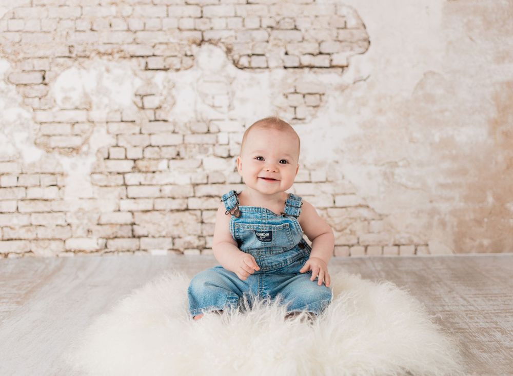 A smiling baby in denim overalls sits on a fluffy white rug against a rustic white brick wall.