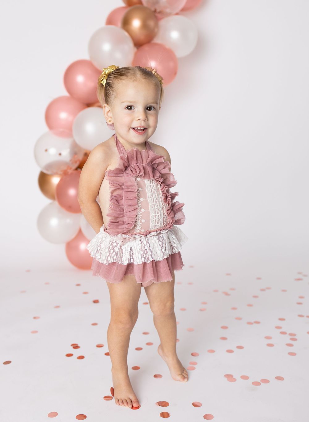 A toddler girl in a pink ruffled lace romper stands barefoot among confetti near rose gold balloons.