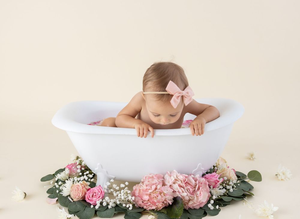 Baby girl in pink bow headband sits in white mini bathtub surrounded by pink roses and hydrangeas on cream background.