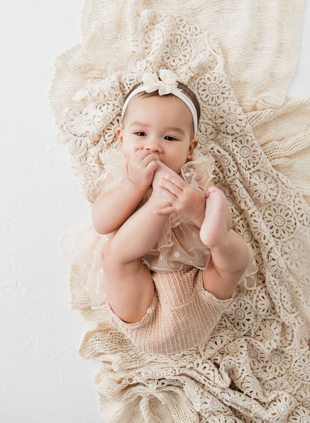 A cute baby in a beige romper and bow headband lies on a cream lace blanket, smiling and holding their feet.