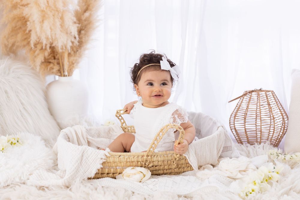 Adorable baby girl in white outfit sitting in wicker basket surrounded by boho-style decor and pampas grass.