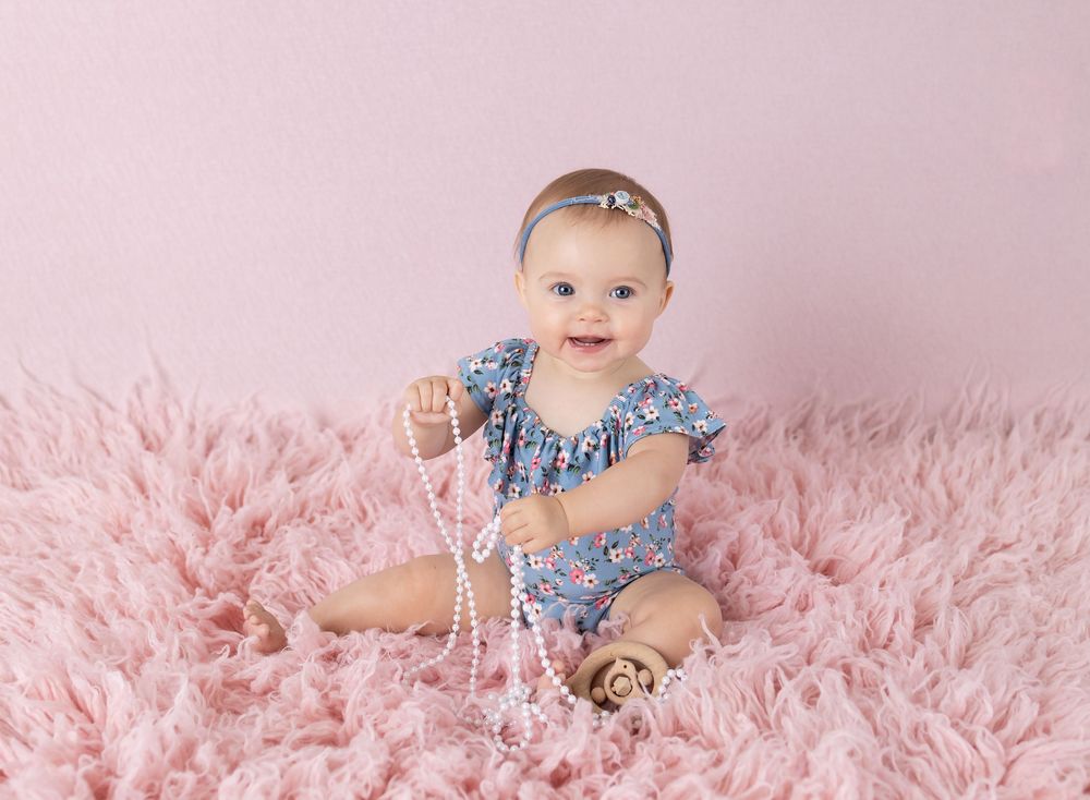 Smiling baby girl in floral romper sitting on pink fluffy rug holding pearl necklace with pink backdrop.