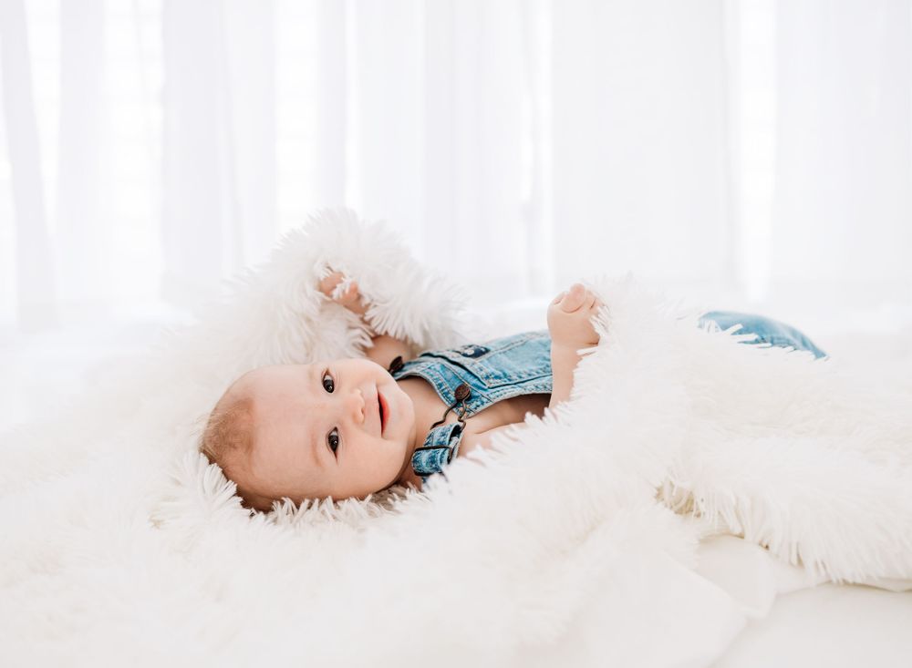 A happy baby in denim overalls lying on a fluffy white blanket near a bright window.