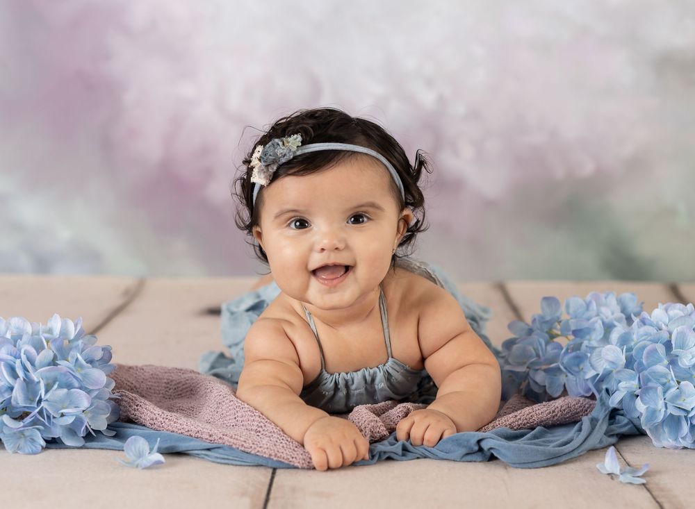 Smiling baby girl in blue dress with floral headband surrounded by blue hydrangeas on wooden floor.