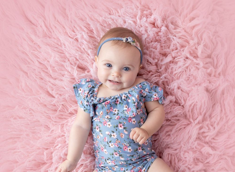 Smiling baby girl in floral blue romper and headband lying on a pink fluffy fur rug.