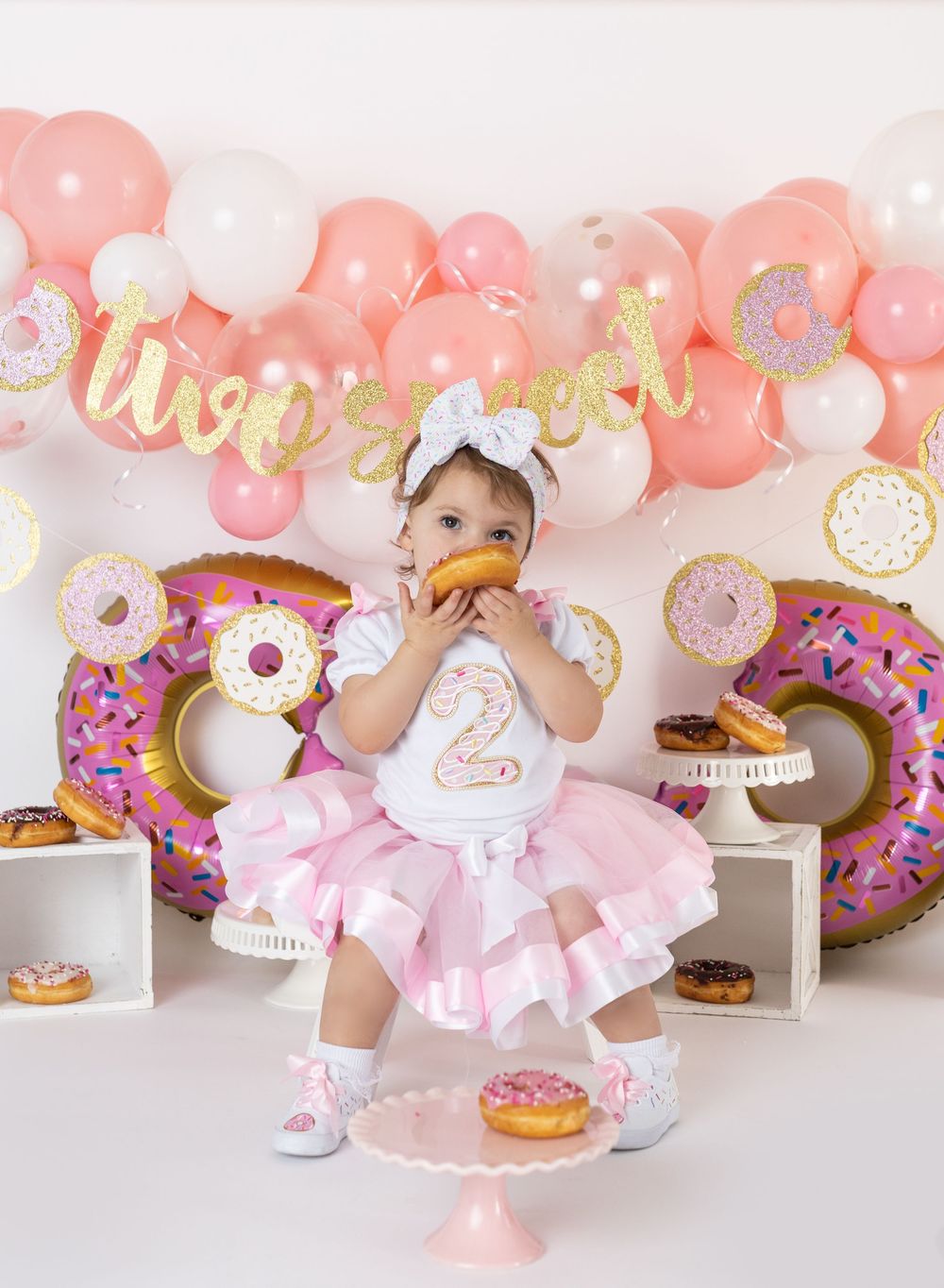 Baby girl in pink tutu celebrating 2nd birthday with donut theme, balloons, and banner at cake smash photo shoot.