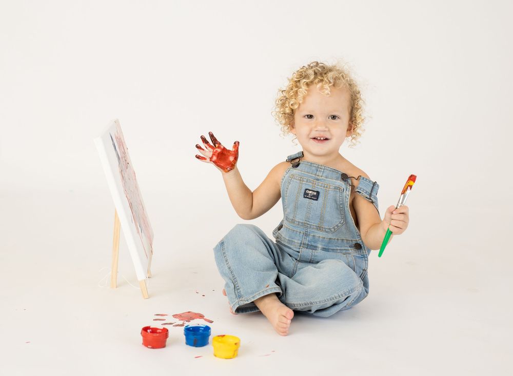 Toddler in denim overalls painting with red paint on hands, holding brush near colorful paint jars on white background.